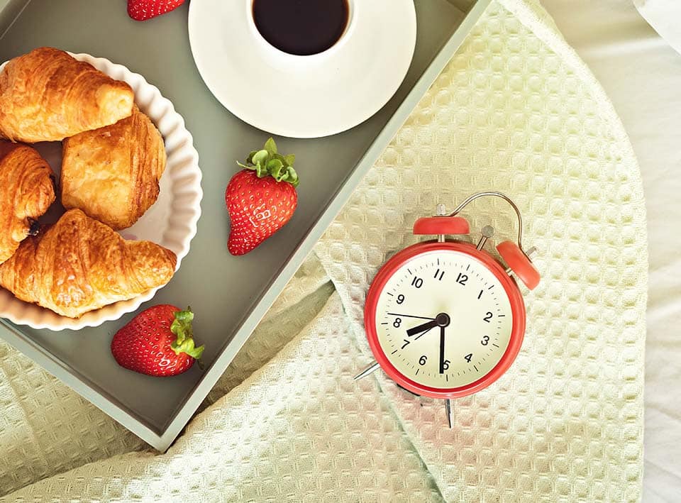 Alarm clock and coffee cup at bed in sunny room. Easy morning start, positive day beginning, waking up, new day, breakfast in bed concept. Flat lay, top view