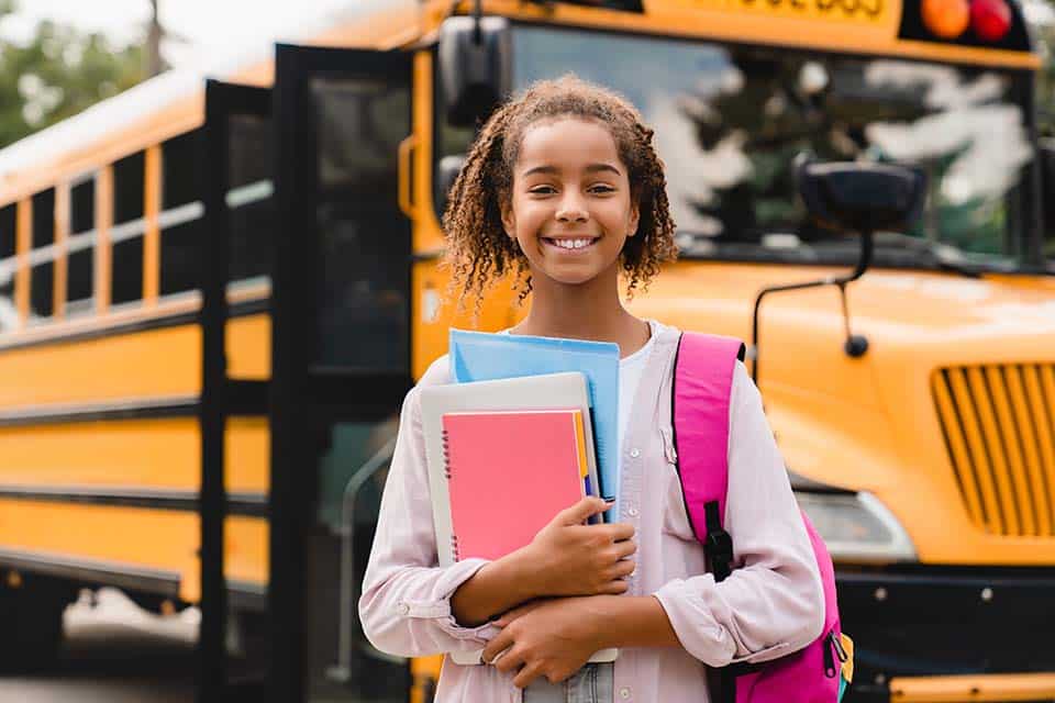 Smiling african-american schoolgirl going back to school with books and copybooks waiting for school bus. New academic year semester.