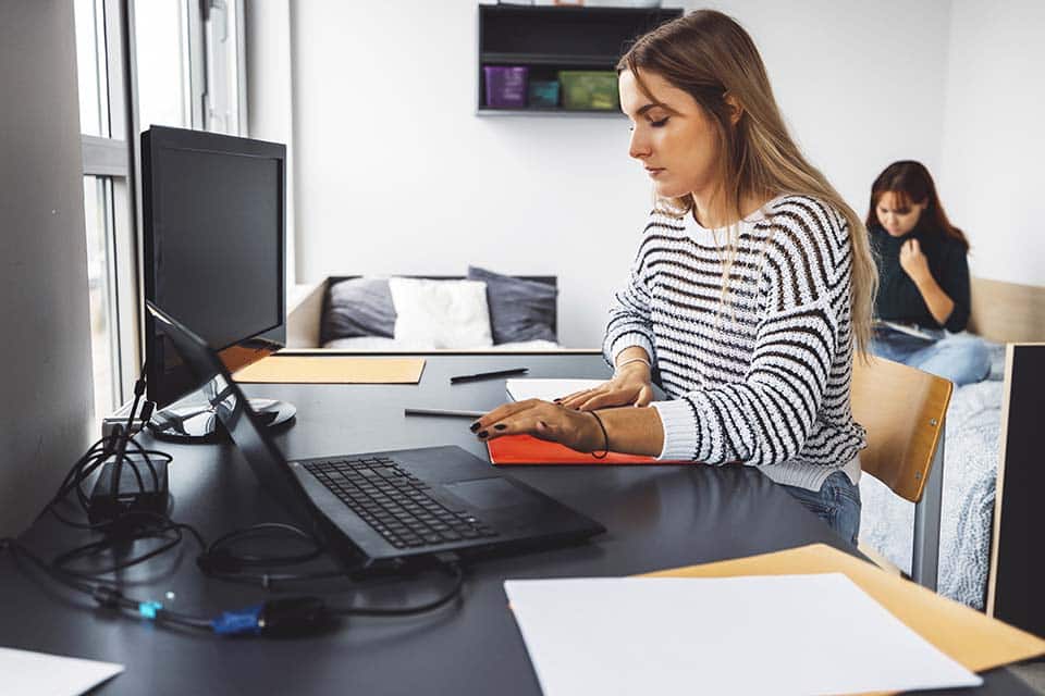 A young woman is sitting in a small dorm room at the community desk working on documents and a computer. You can see her roommate sitting in the background on one of the two the twin sized mattress in the room